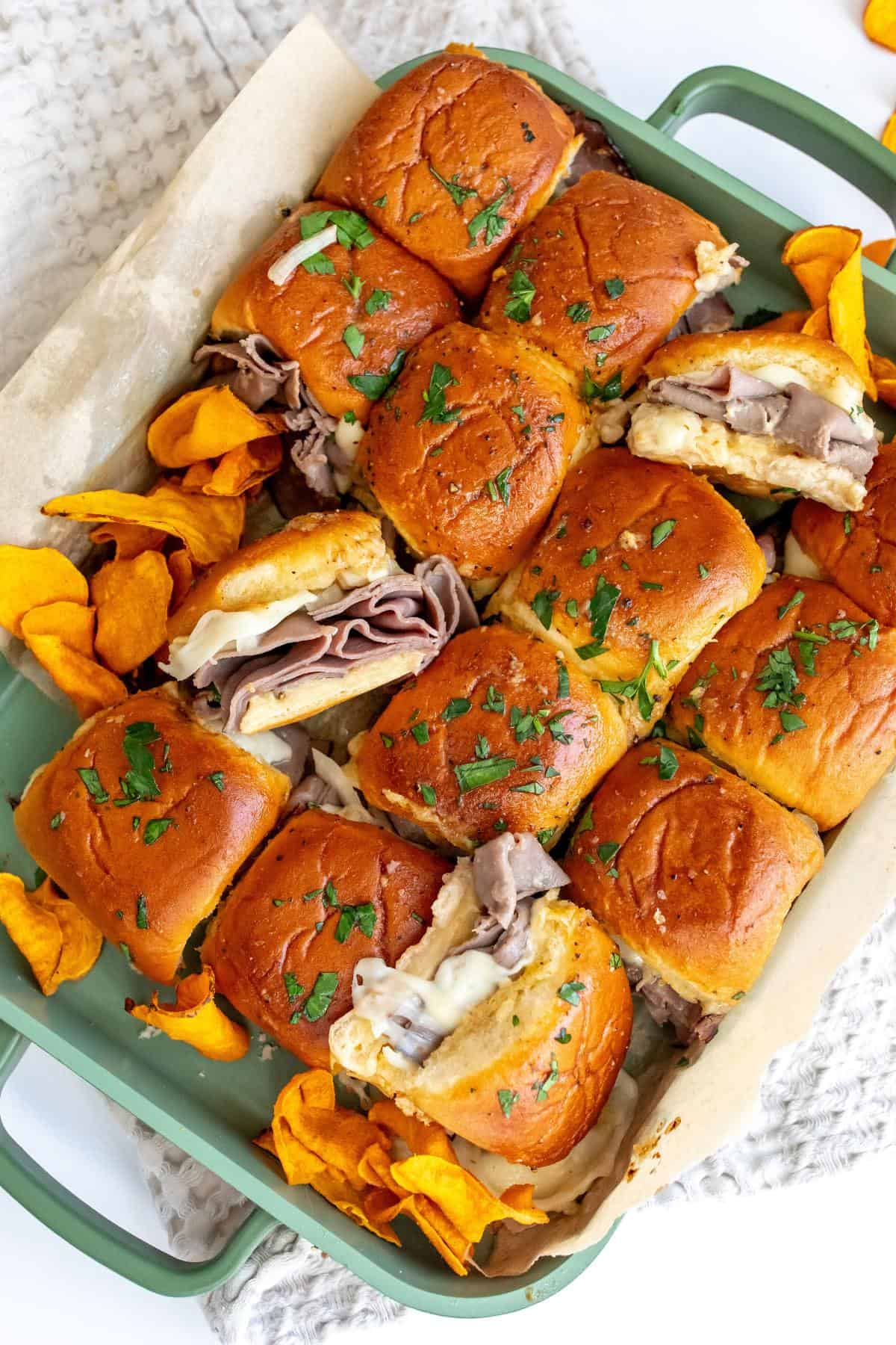 Roast beef sliders in a green pan with sweet potato chips on the side. 