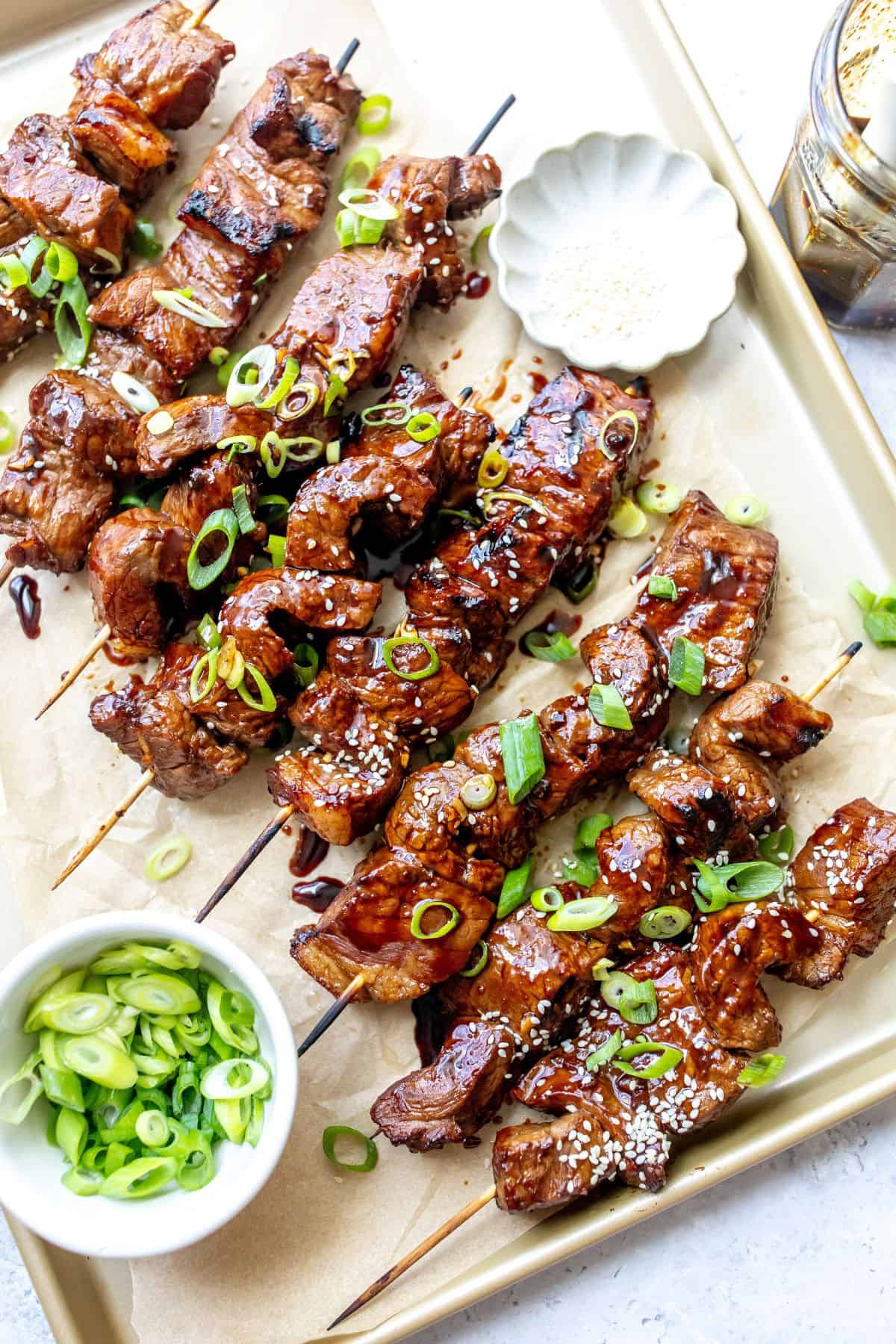 A baking sheet with steak skewers on them with sauce and green onions.