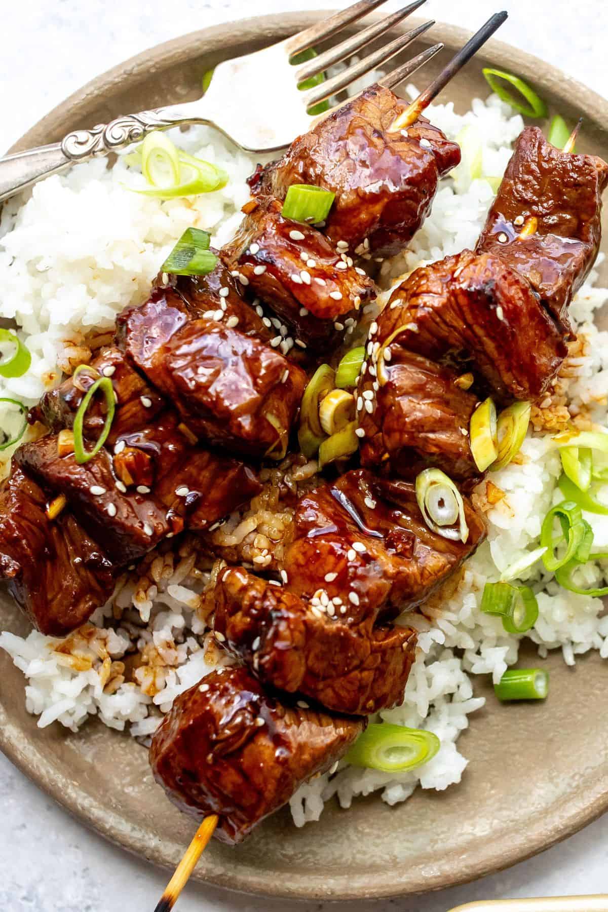 Grey plate with steak kabobs laying on a bed of rice with green onions.