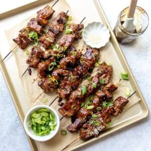 A baking sheet with steak skewers on them with sauce and green onions.