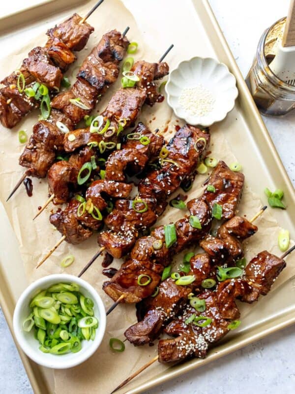 A baking sheet with steak skewers on them with sauce and green onions.