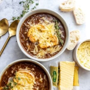 Two bowls with healthy french onion soup in them, gold spoons off to the side and cheddar shredded.