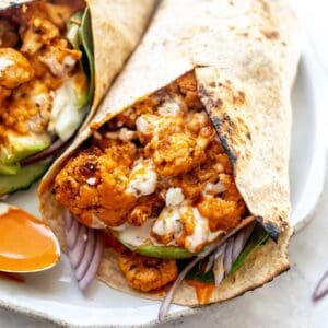 Buffalo cauliflower wraps&nbsp;on a white plate with a spoon to the side for serving sauce.
