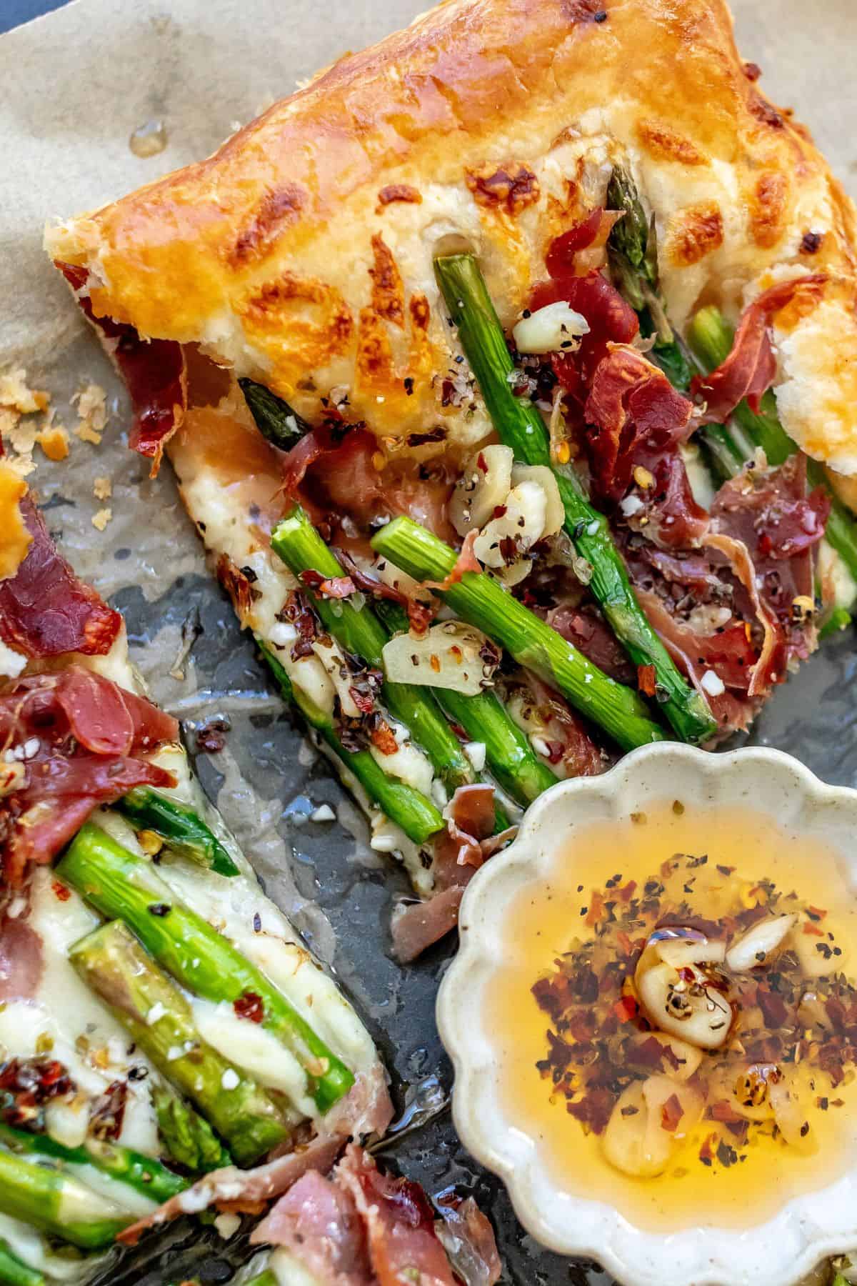 Asparagus puff pastry with prosciutto on it and a spoon to the side with oil and garlic. 