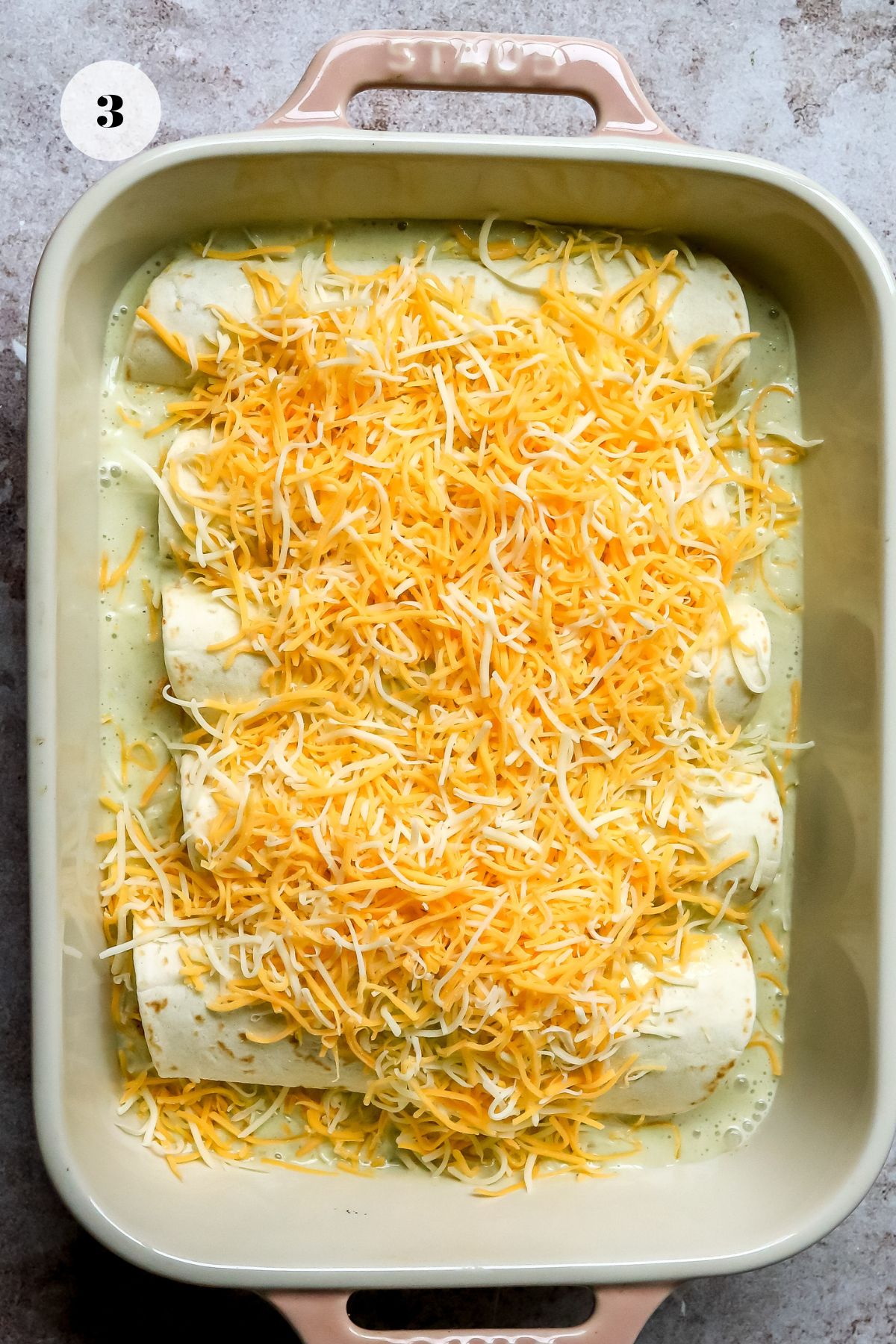 Enchiladas in a pink baking dish with lots of shredded cheeses on top before going into the oven.