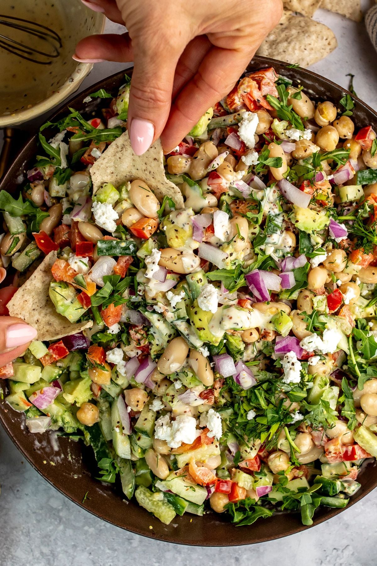 White bean salad in brown bowl with a hand dipping a tortilla chip into it.