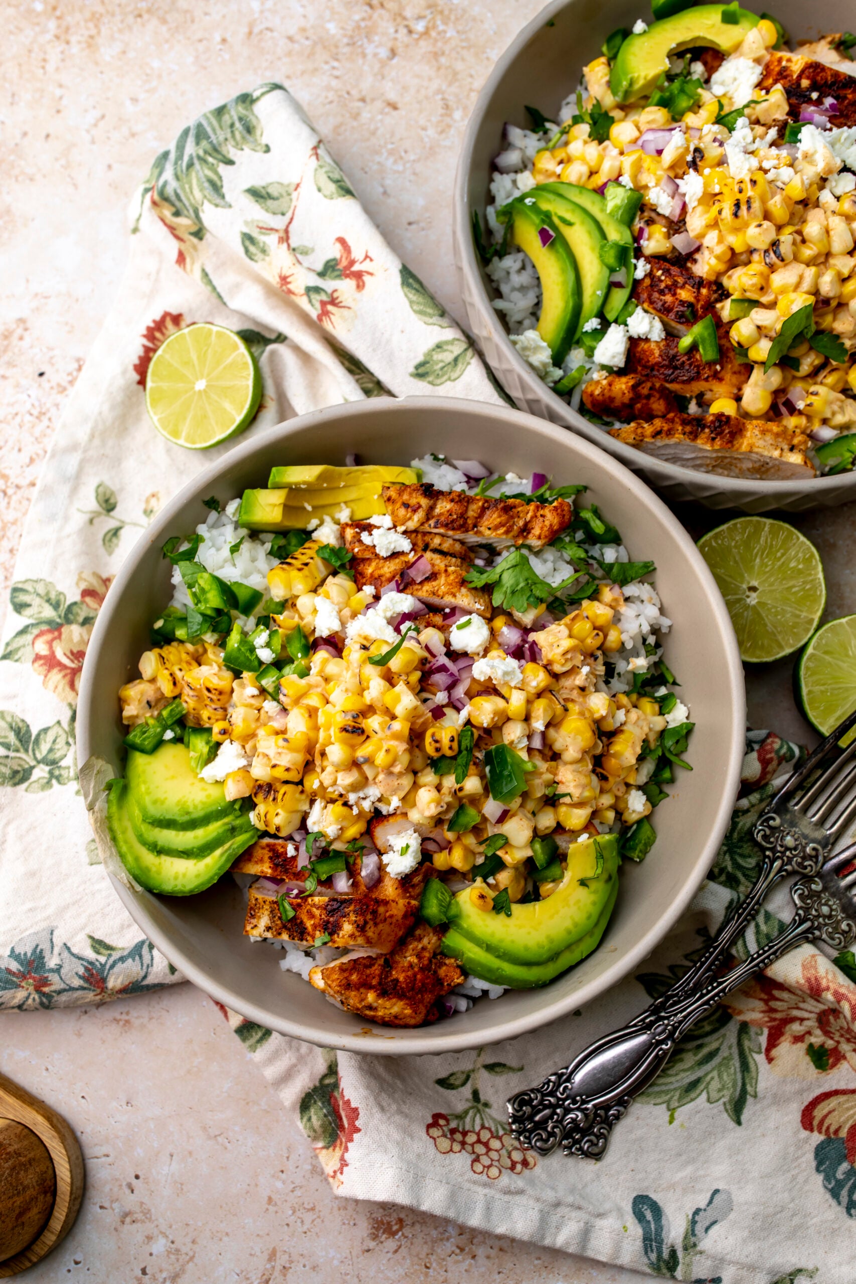 Two Chicken Street Corn Rice Bowls on a counter with a floral napkin underneath and silver forks to the side.