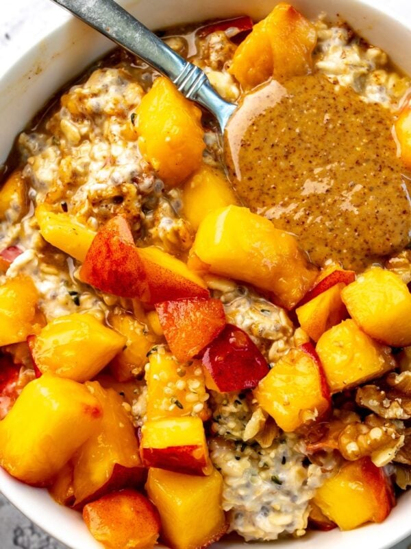 A spoon with almond butter on it rests in a bowl of peach overnight oats.