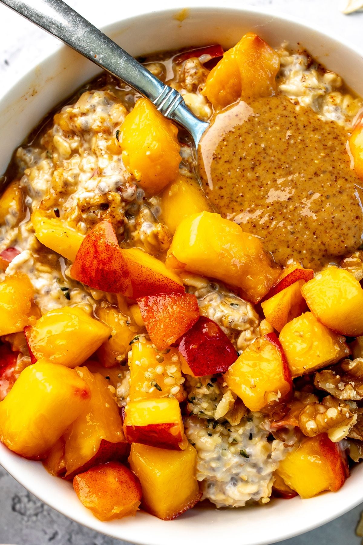 A spoon with almond butter on it rests in a bowl of peach overnight oats.