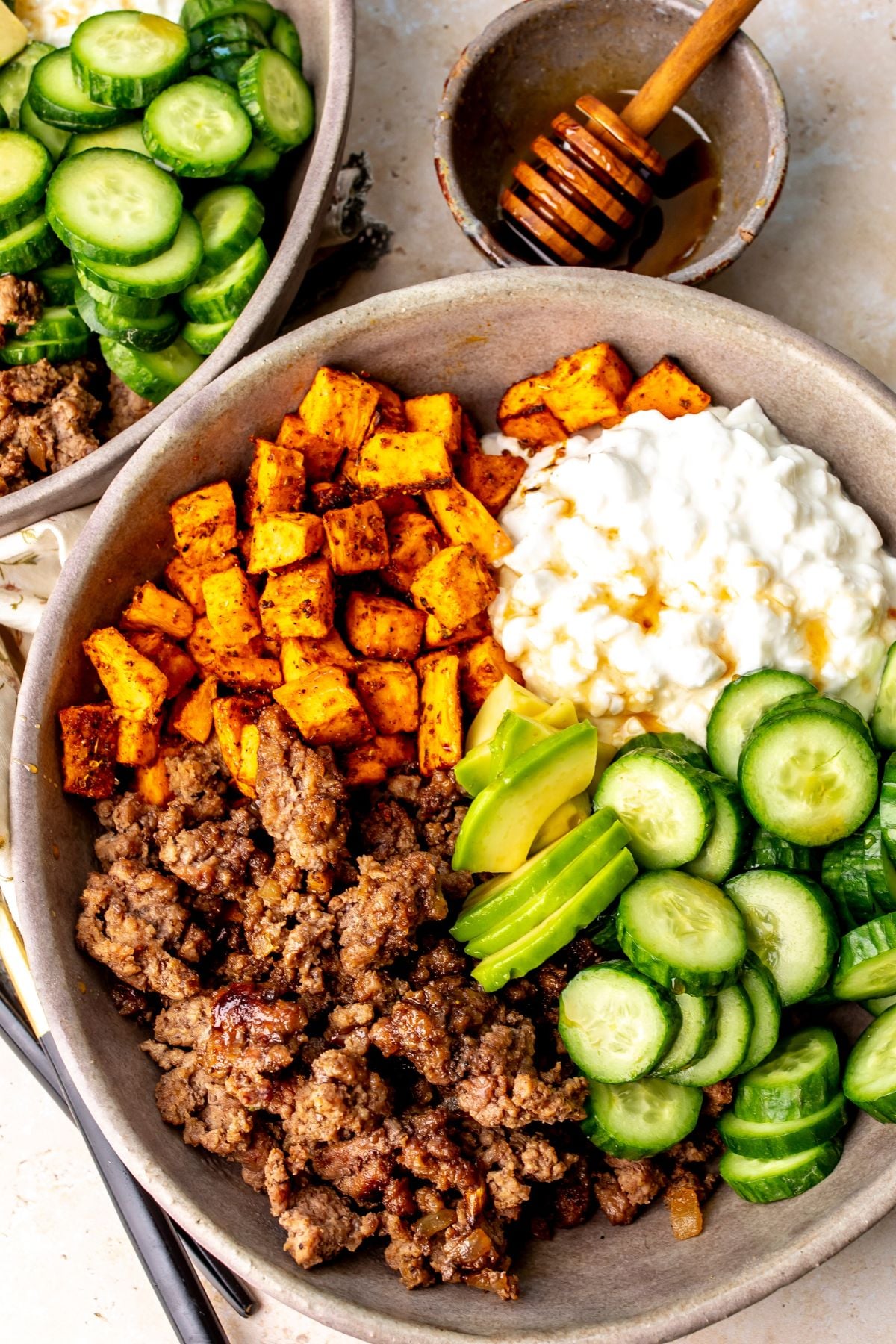 Hot honey ground beef bowls with sweet potatoes, avocado, cucumbers, and cottage cheese in a bowl on a countertop.