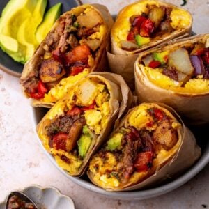 Steak breakfast burritos in a bowl on a countertop next to a bowl of sliced avocado.