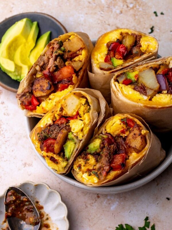 Steak breakfast burritos in a bowl on a countertop next to a bowl of sliced avocado.