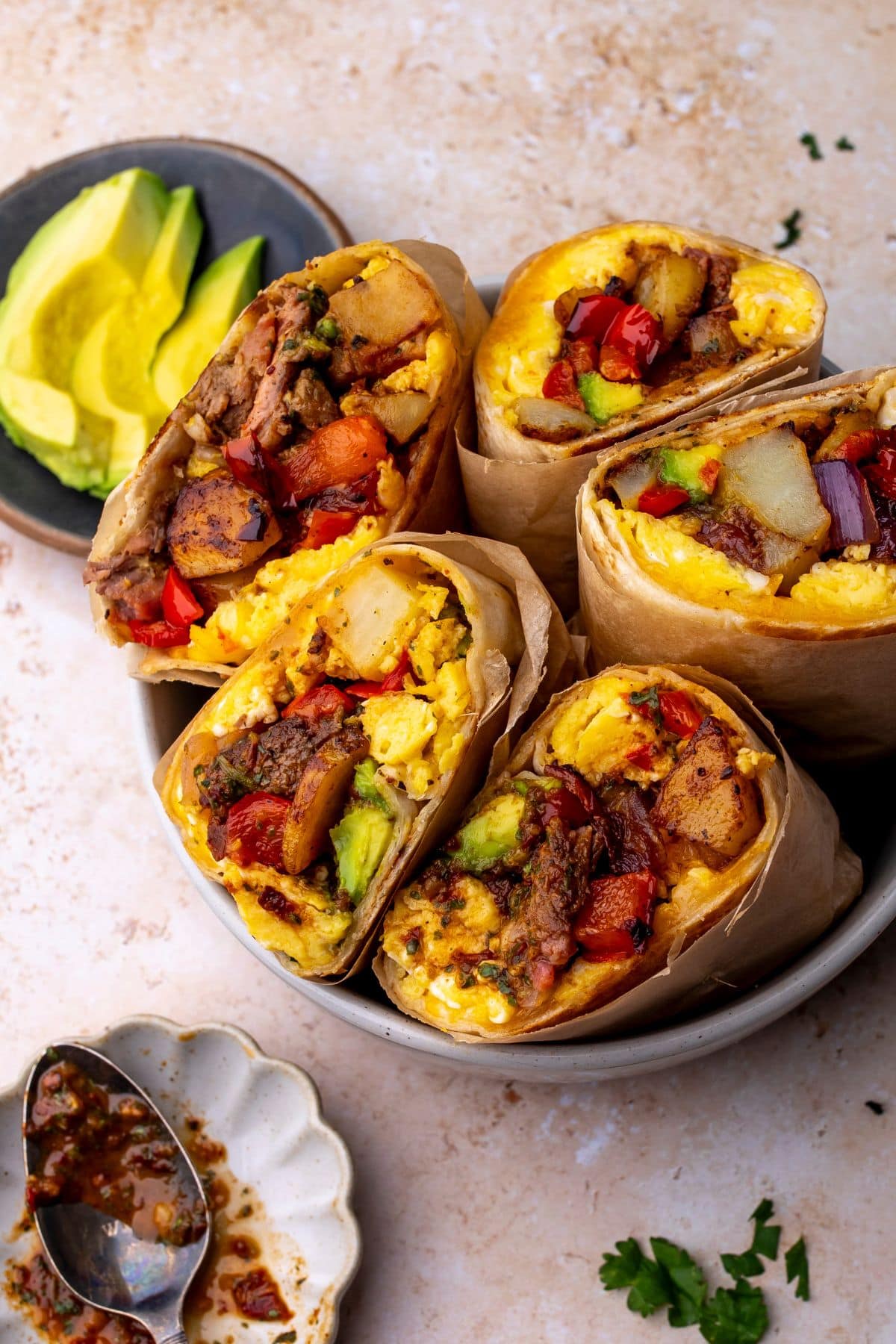 Steak breakfast burritos in a bowl on a countertop next to a bowl of sliced avocado.