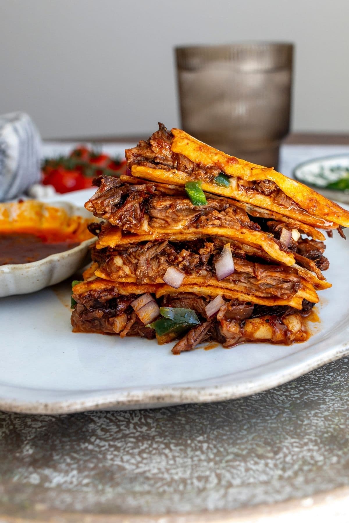 Birria quesadilla sliced into four large pieces and stacked on a plate with a bowl of the extra comsomme to the side.