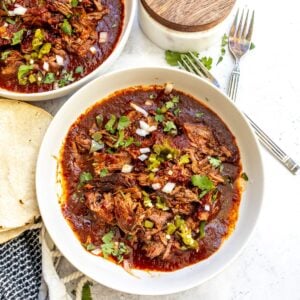 Slow cooker birria in a white bowl on a countertop garnished with chopped cilantro and onion.