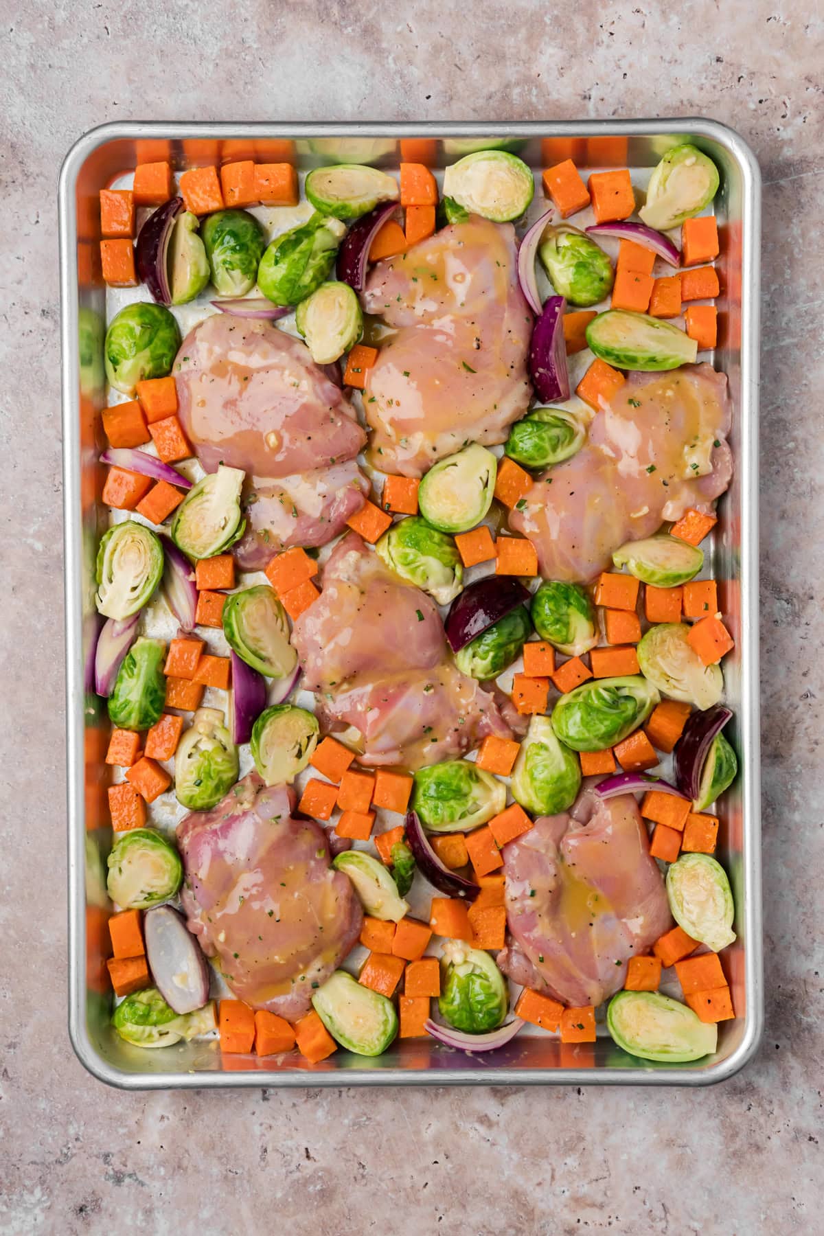 The chicken thighs with the brussels sprouts and sweet potatoes on the baking sheet before baking in the oven.