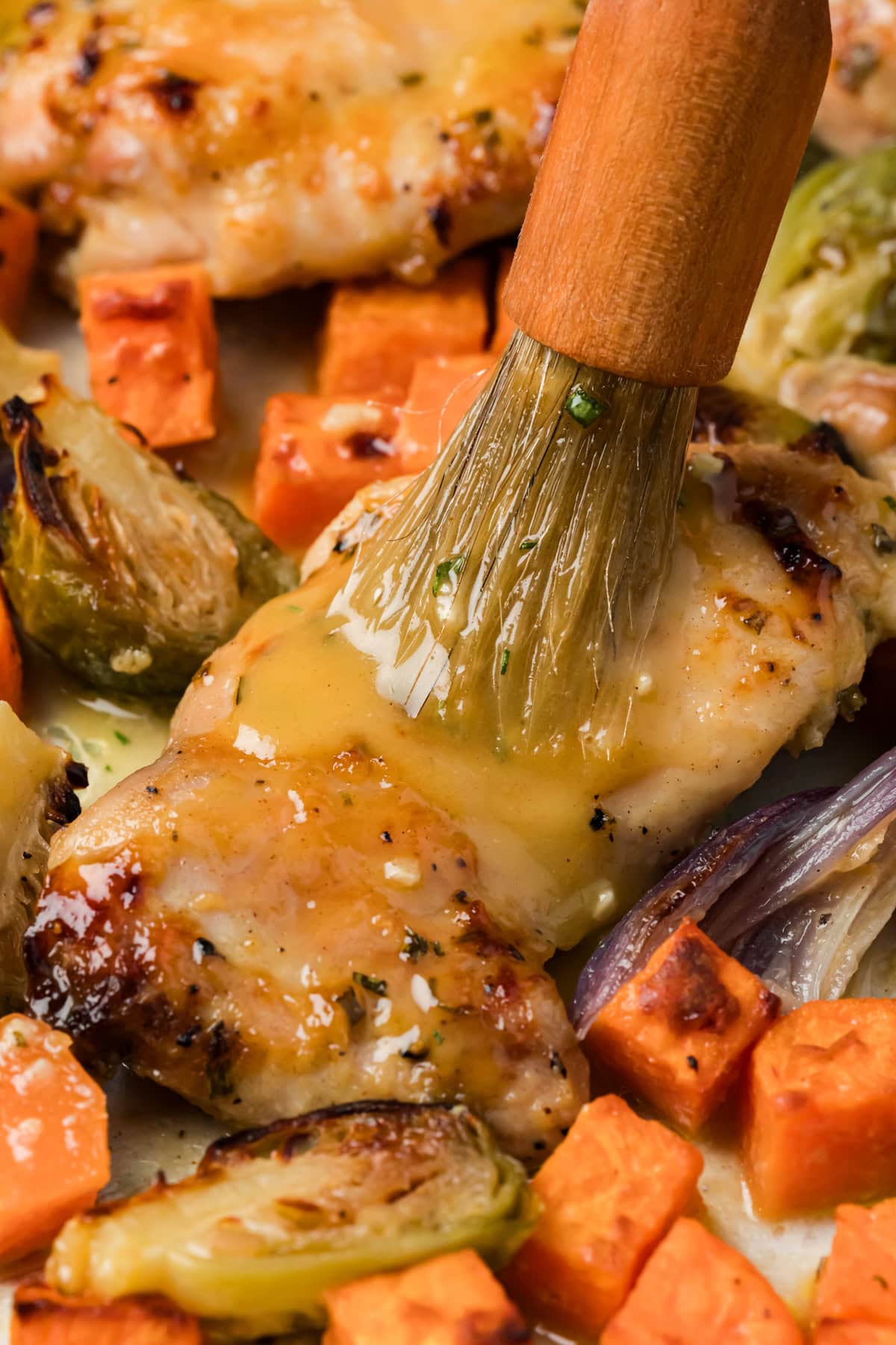 A pastry brush brushes the reserved maple Dijon sauce onto a chicken thigh on the sheet pan.